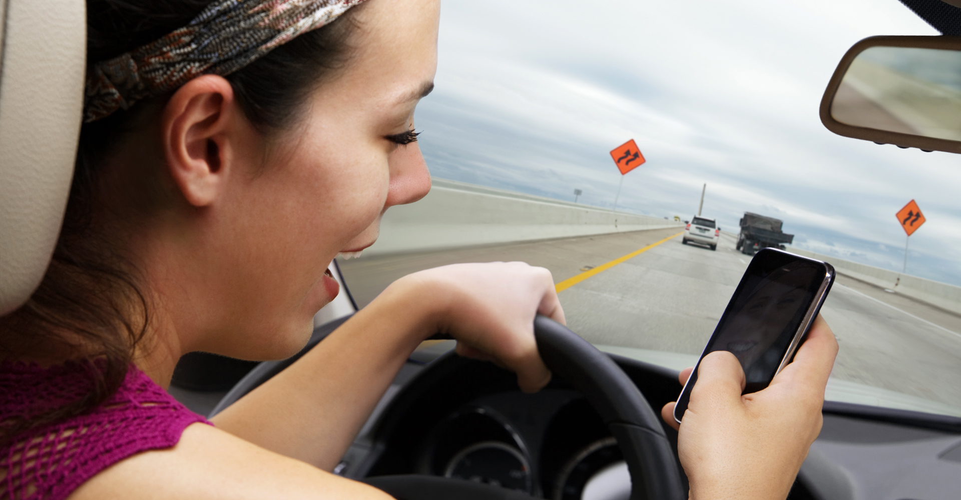 young woman driving on highway while reading / writing text on smart phone.